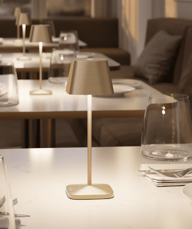 A modern table lamp with a gold base and shade sits on a set dining table in a well-lit restaurant, surrounded by plates, glasses, and silverware.